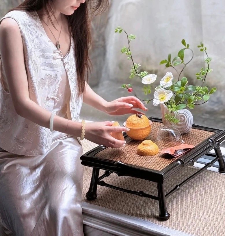 Young woman setting up a natural bamboo small tea table with a vase of flowers and fruits, placed on a tatami mat in a serene home environment.