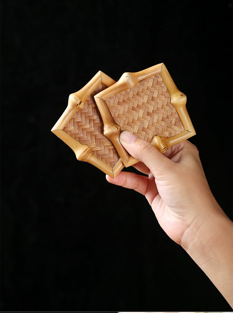 A hand displaying two decorative square, woven bamboo coasters from the 10pcs Bamboo Tea Coasters set against a black background.