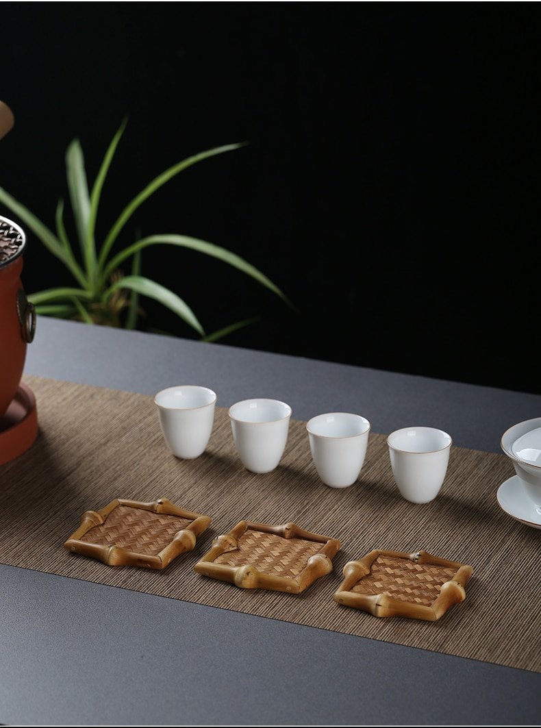 On a dark table, a potted plant complements a minimalist and serene tea setup featuring four small white porcelain cups and bamboo coasters from the "10pcs Bamboo Tea Coasters - Tea Ceremony Accessories, Kitchen Supplies" collection, all elegantly arranged on a woven mat. The black background accentuates the tranquil ambiance.