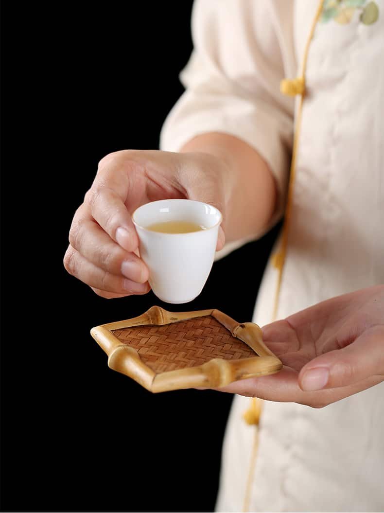 A person wearing a light-colored traditional outfit holds a small white teacup filled with tea in one hand and one of the 10pcs Bamboo Tea Coasters - Tea Ceremony Accessories, Kitchen Supplies in the other, set against a dark background.