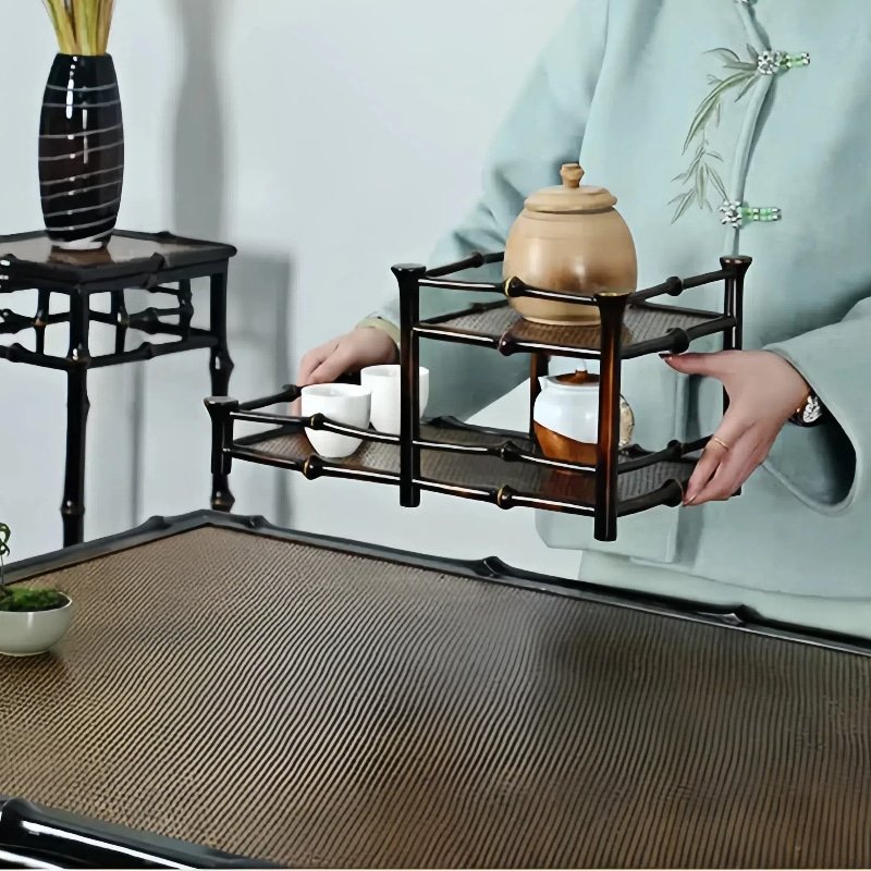 A person in a light green traditional outfit is holding a 2-Tier Qiong Bamboo Tea Shelf, which serves as both a teaware organizer and display stand, featuring a teapot and cups. The setting includes a table with a brown woven surface, and in the background, there's a small table adorned with tall grasses in a vase.