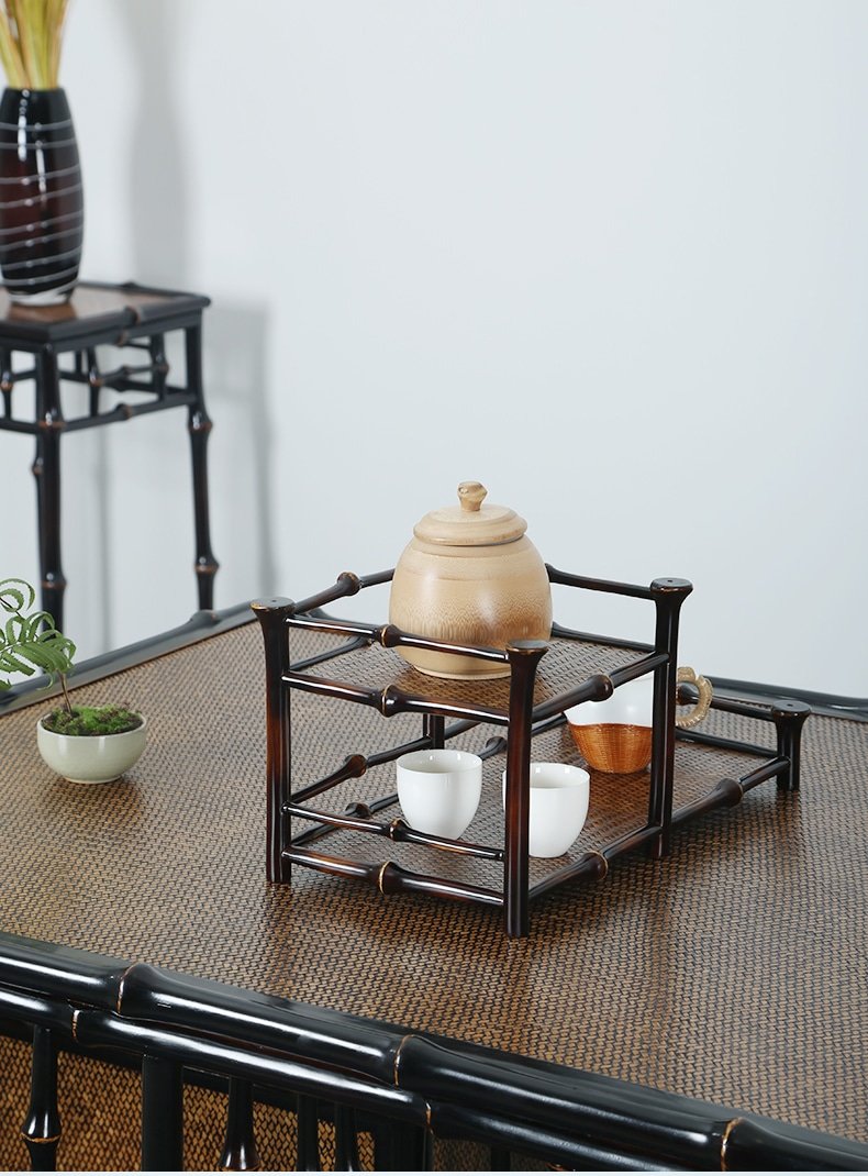 The 2-Tier Qiong Bamboo Tea Shelf serves as a wooden tray ensemble featuring a teapot, two white teacups, and a small wooden bowl, complemented by a potted plant on the side. In the background stands a dark wooden display with a tall vase.
