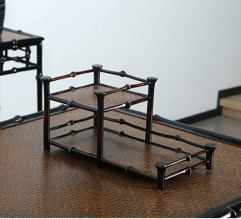 A compact, two-tiered 2-Tier Qiong Bamboo Tea Shelf rests on a textured brown surface. The shelf features open sides and is crafted from dark bamboo rods, showcasing a simple and elegant design ideal for organizing and displaying teaware.