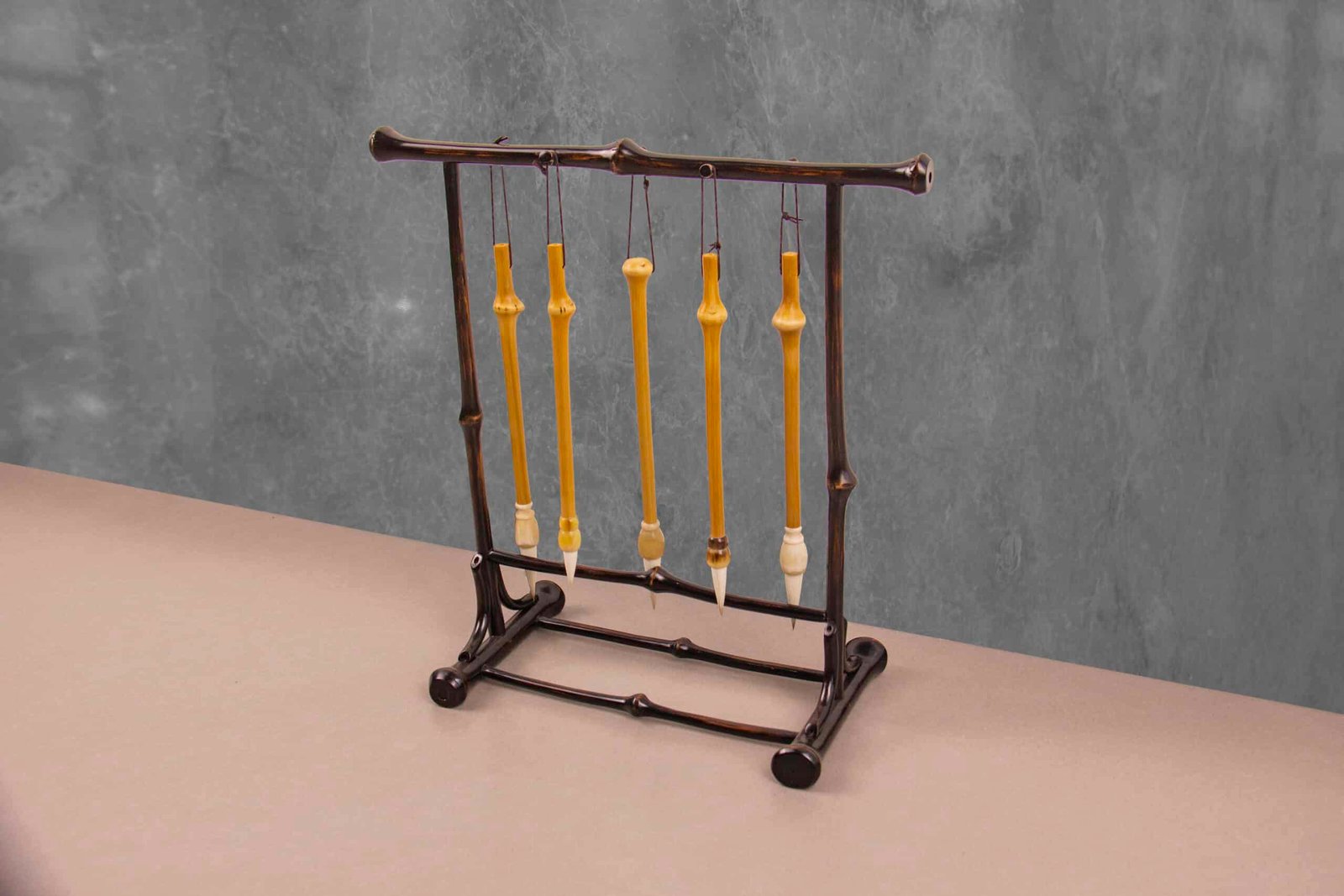 A Chinese Calligraphy Brush Pen Rack with 5 Hooks from Qiongzhu Bamboo elegantly graces a dark wooden stand against a gray textured background. The bobbins hang vertically, featuring intricate designs that evoke the artistry of Chinese calligraphy. The stand showcases a sturdy, ornate base.