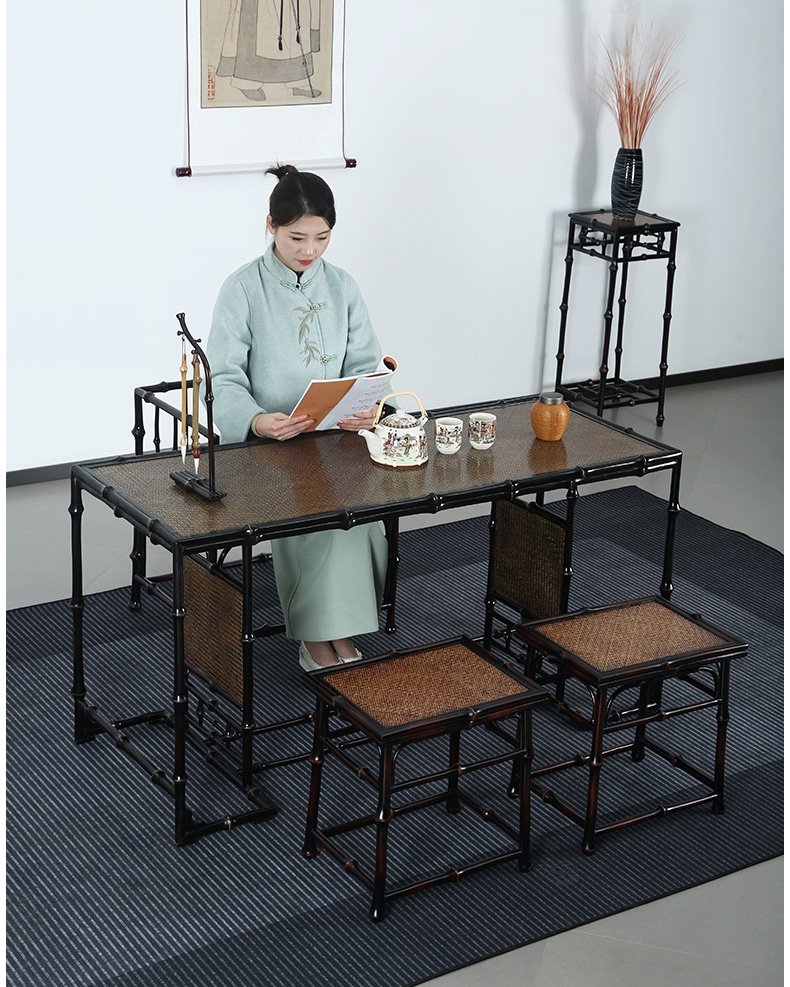 A woman in traditional attire is seated at a Handmade Chinese Qiongzhu Furniture Bamboo Table for Tea, immersed in her book. The table, with its bamboo-style design, displays a teapot and mugs. Nearby, a matching chair and stand with a vase enhance the minimalist decor, complemented by an elegant wall hanging.