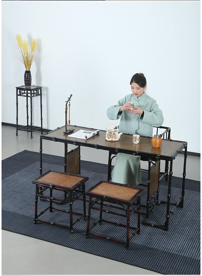 A woman in a green robe sits at the Handmade Chinese Qiongzhu Furniture bamboo tea table, preparing tea. The set includes two small stools and an orange teapot. In the background, a black vase with yellow flowers stands gracefully, complementing the elegance of the handmade furniture.