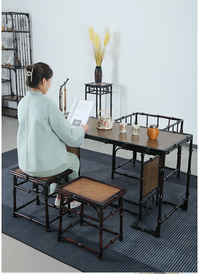 A person with a bun hairstyle, wearing a light blue sweater, sits at a Handmade Chinese Qiongzhu Furniture: Bamboo Table for Tea, Home Use, reading a book. The table holds a teapot and cups, while a vase with yellow flowers rests on a nearby stand. The minimalist setting exudes calm and tranquility.