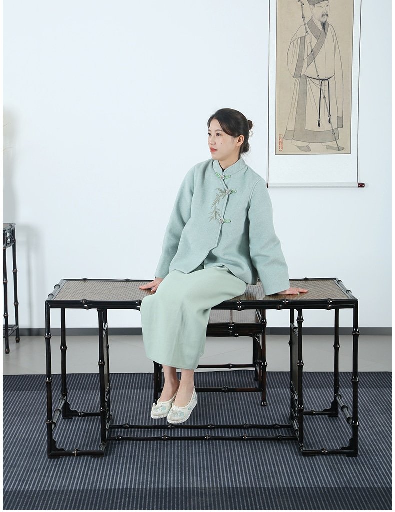 A woman in a light blue traditional outfit sits on a woven bench beside the Handmade Chinese Qiongzhu Furniture: Bamboo Table for Tea, arranged in a minimalist room. A framed artwork of a historical figure graces the wall behind her, and a black and grey striped rug adorns the floor, enhancing the serene ambiance.