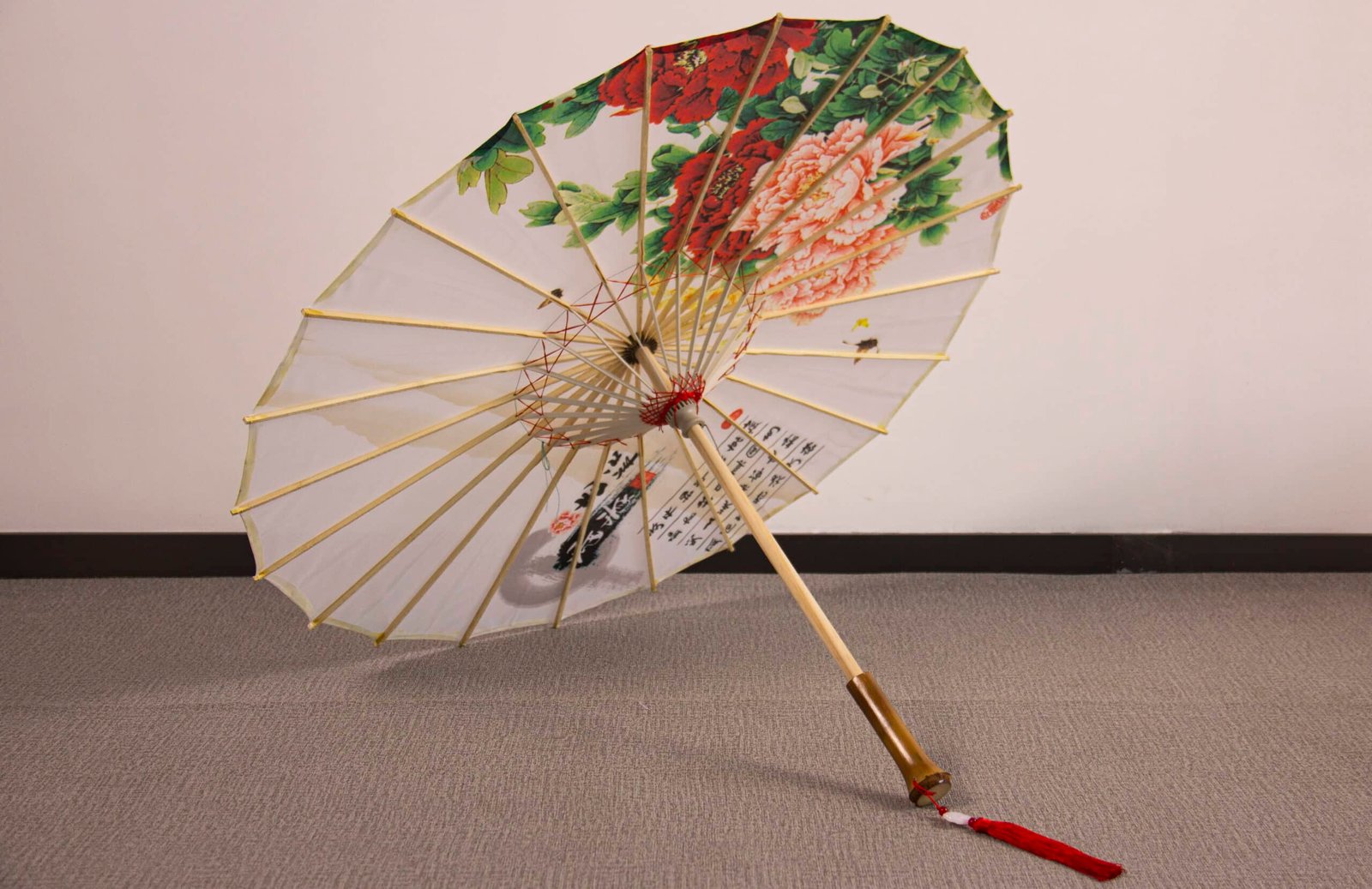 An Oil Paper Umbrella - Decorative & Antique Handmade & Waterproof Umbrella with a wooden handle is open and resting on the floor. The umbrella showcases vibrant floral patterns in red and green, accented with calligraphy symbols on a white background, while a red tassel dangles from the handle.