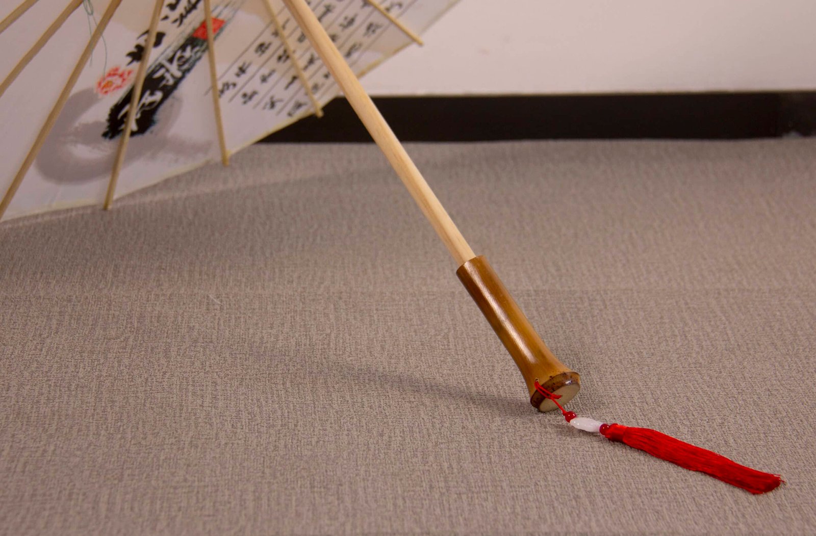 A decorative and antique handmade Oil Paper Umbrella rests open on a grey carpeted floor. It showcases delicate floral patterns and calligraphy, with a wooden handle that features a red tassel extending from it.