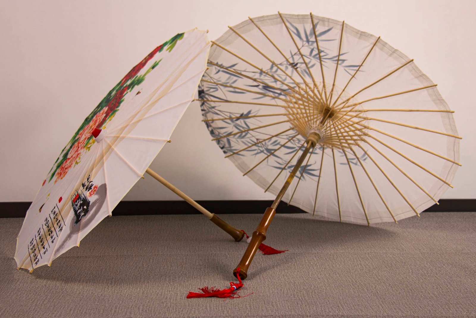 A pair of Oil Paper Umbrellas, featuring traditional wooden handles, are open and leaning against each other. One umbrella showcases a floral design, while the other is adorned with bamboo motifs. They are displayed on a gray carpeted floor against a white wall.