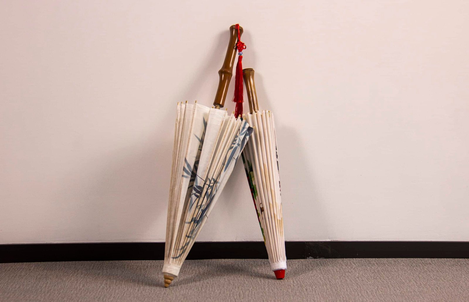 Two closed Oil Paper Umbrellas with bamboo handles stand against a white wall on a gray carpeted floor. The umbrellas are embellished with decorative patterns on their fabric.