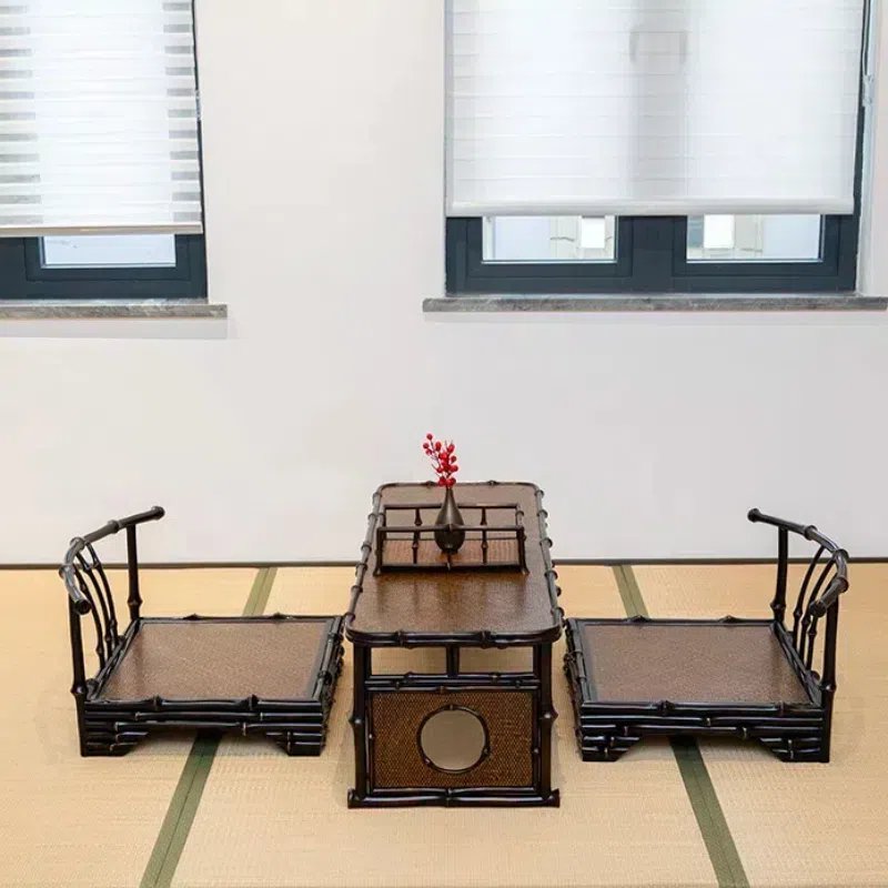 A minimalist room with a bamboo tea table set, featuring two chairs and a low table. The table holds a small vase with red flowers. The room has tatami mat flooring and two windows with blinds.