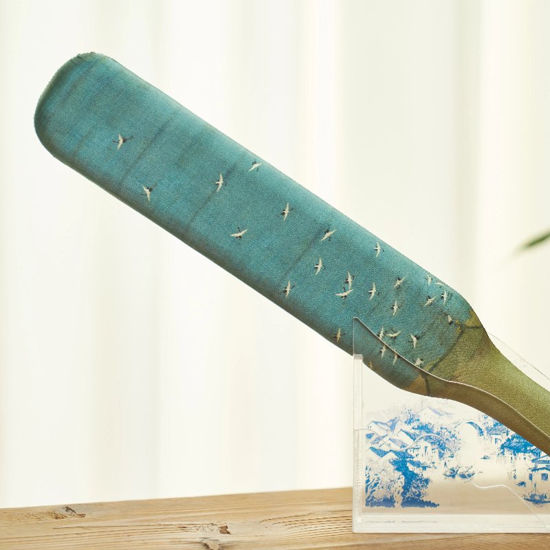 An artistic wooden paddle with a gradient blue hue and small white bird motifs sits in a transparent stand on a wooden surface, set against a blurred light background.
