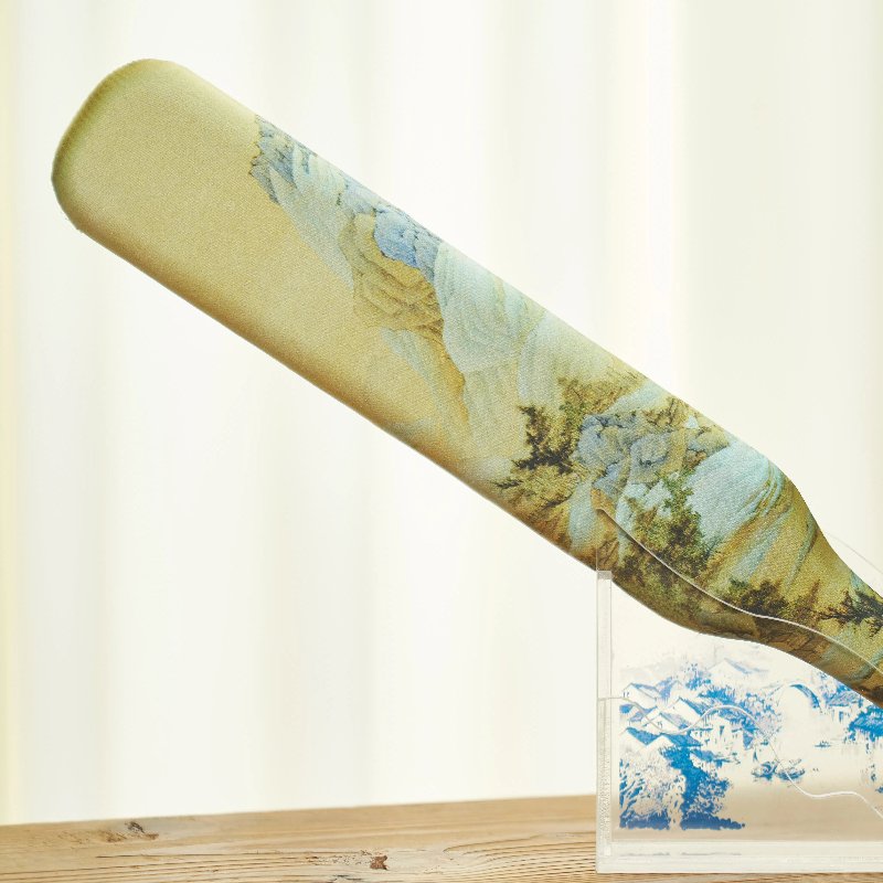 A decorative cricket bat with a landscape painting stands upright on a wooden surface. The design features subtle colors and intricate nature scenes, with a clear stand supporting it from below. A light, blurred background is visible.