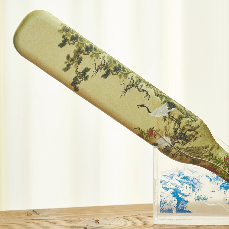 A decorative paddle with a nature scene featuring cranes and trees, displayed on a clear stand against a blurred light background. The design includes intricate details and delicate colors, evoking a sense of tranquility.