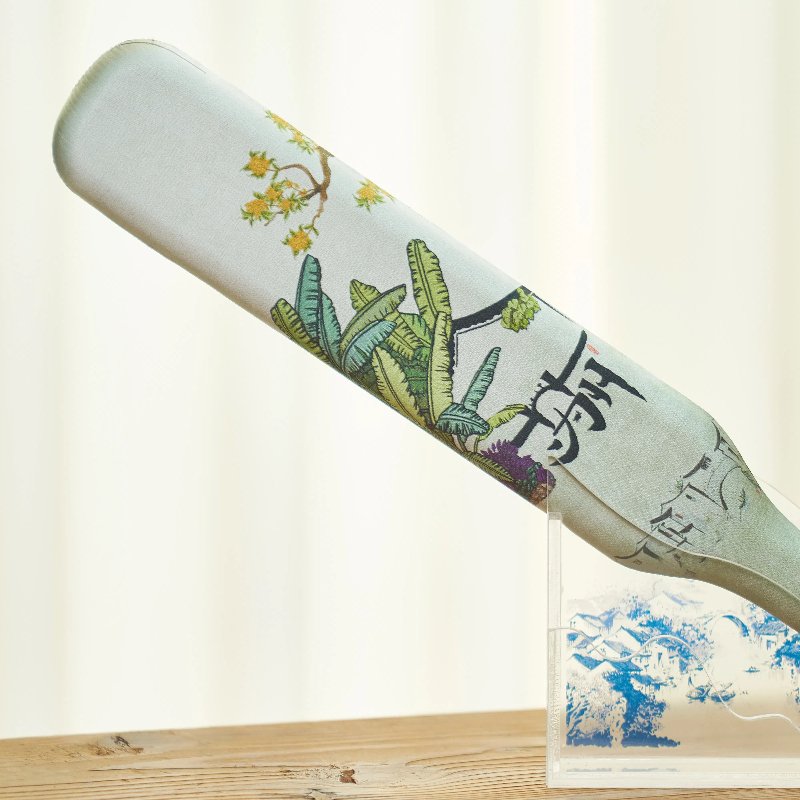 A decorative flat object with botanical motifs and calligraphy stands on a transparent base. The design includes green plants and yellow flowers, set against a soft, light background.