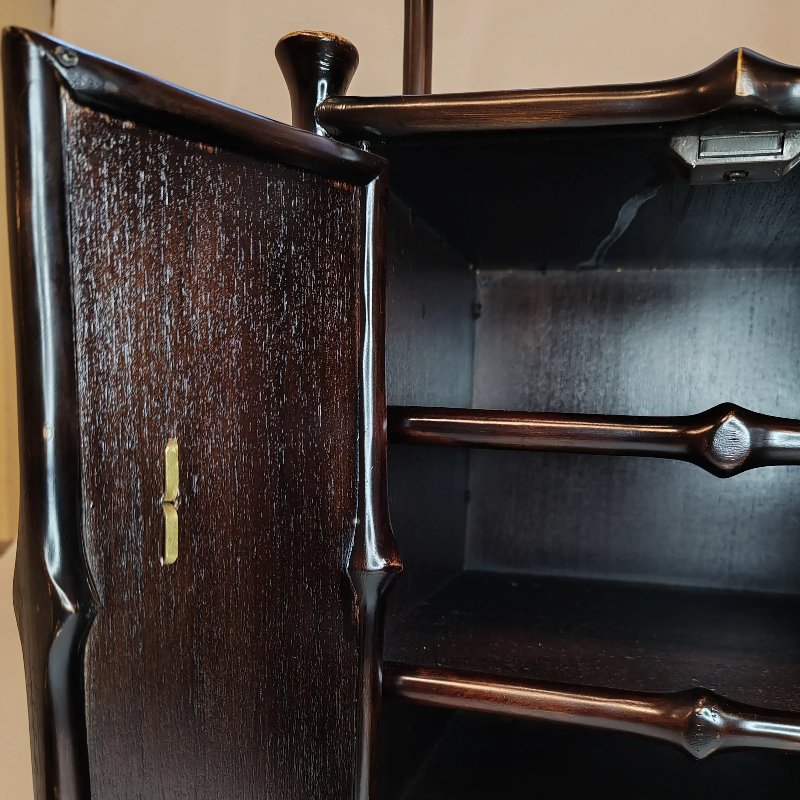 A close-up of an open wooden cabinet with a dark finish. The door is slightly ajar, revealing two shelves inside. The cabinet has a glossy surface and visible grain patterns. There is a small latch on the inside of the door.