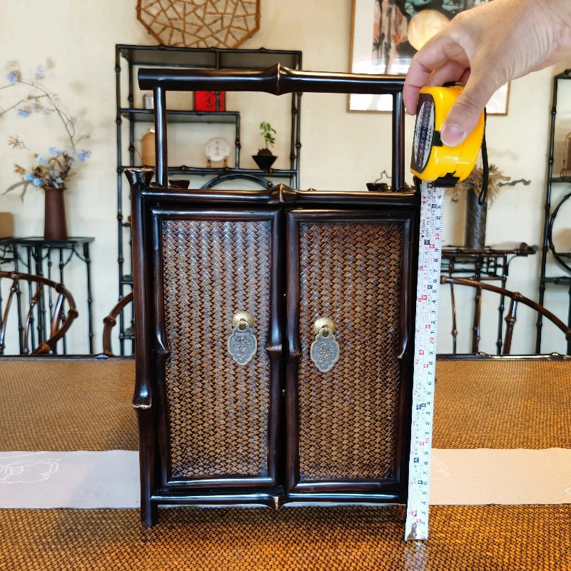 A hand holds a tape measure against a small, ornate wooden cabinet with decorative details. The cabinet, placed on a table, features two doors with metal handles and woven paneling. Background includes wooden chairs and shelves with decorative items.