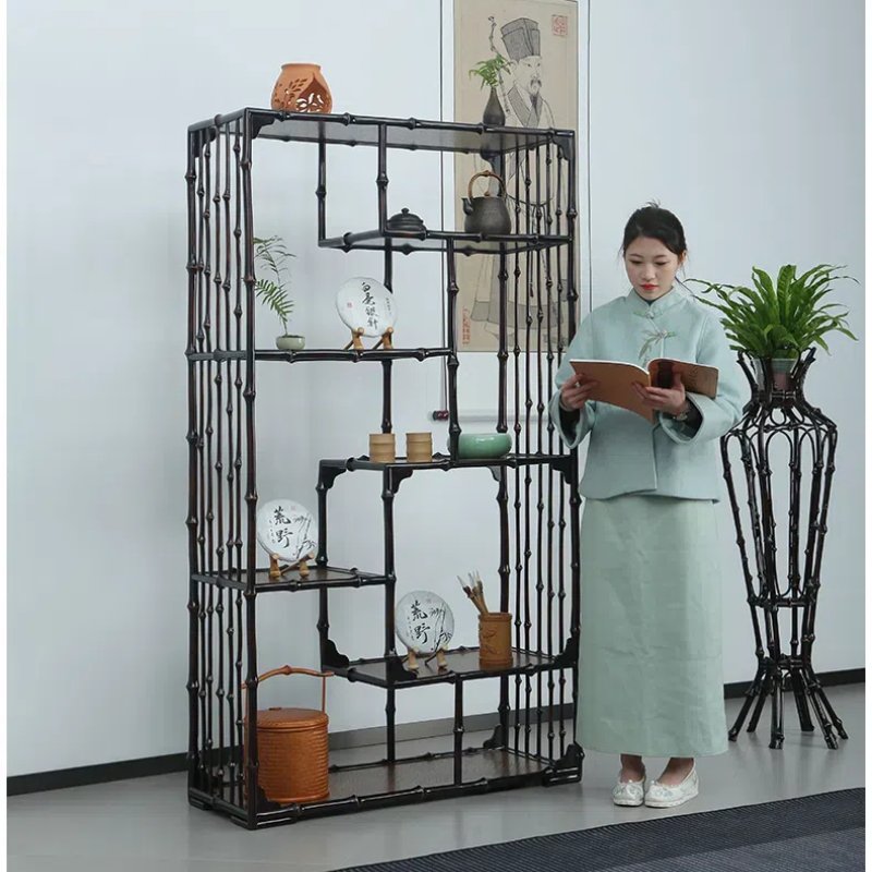 A person in traditional clothing stands reading a book near a tall, black bamboo bookshelf. The shelf displays various decorative items and plants. In the background, theres a hanging scroll with an illustration.