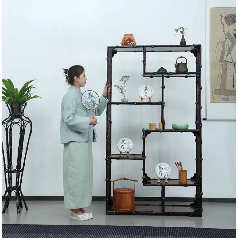 A woman in traditional attire holds a circular fan and arranges items on a decorative shelf. The shelf contains pottery, plants, and decorative plates. A potted plant stands beside her, and a painting hangs on the wall.