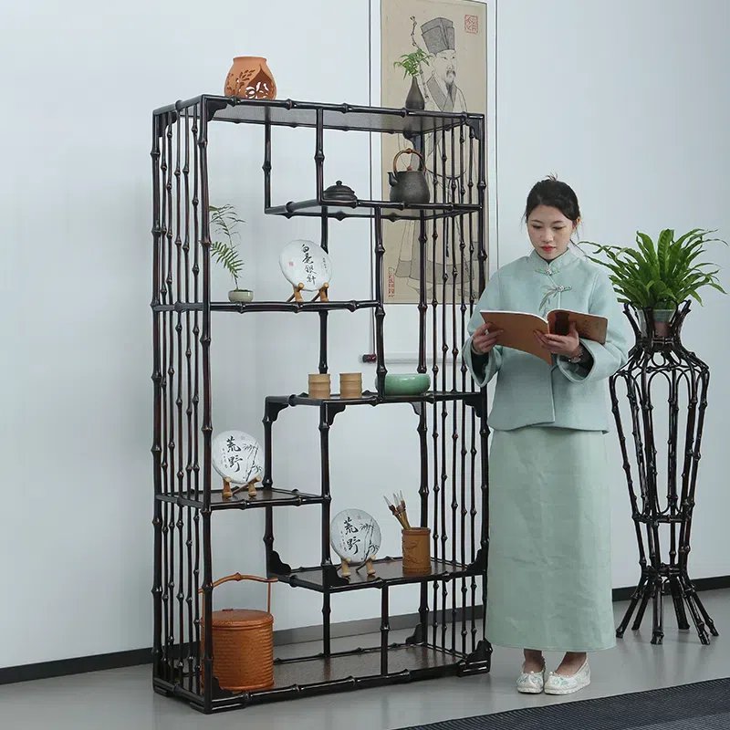 A woman in traditional attire stands reading a book beside a tall, decorative bamboo shelf displaying various pottery and potted plants. The background features a framed portrait hanging on the wall.