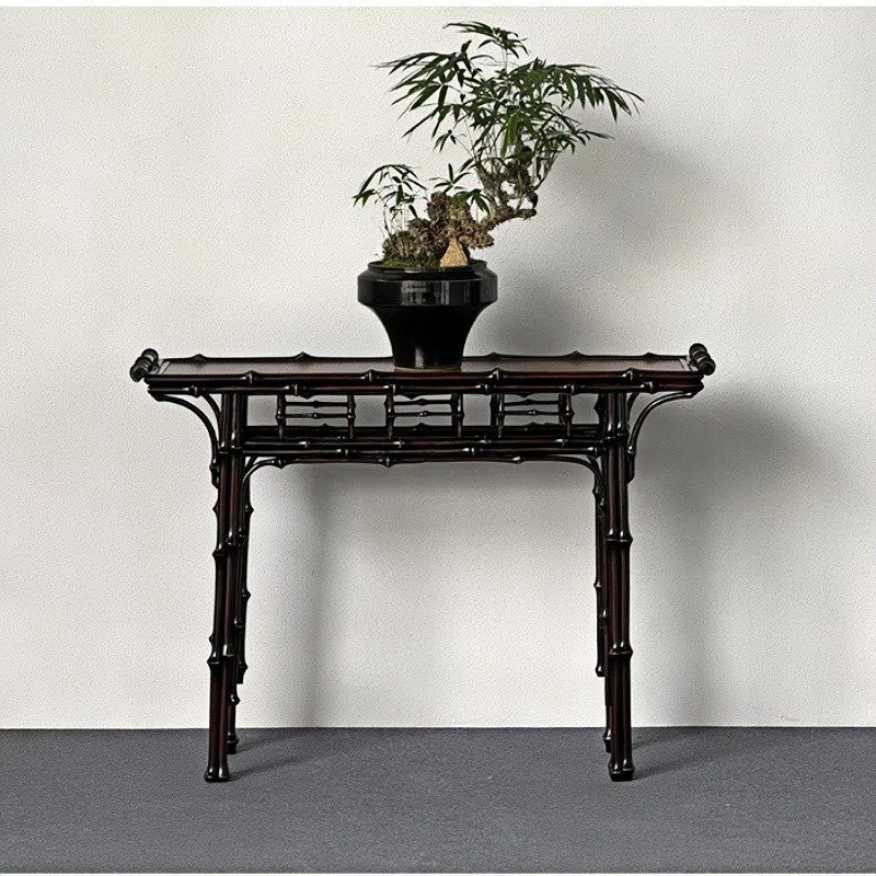 A small bonsai tree in a black pot sits on an elegant, dark wood table with a bamboo-like design. The setup is against a plain white wall and a gray floor.