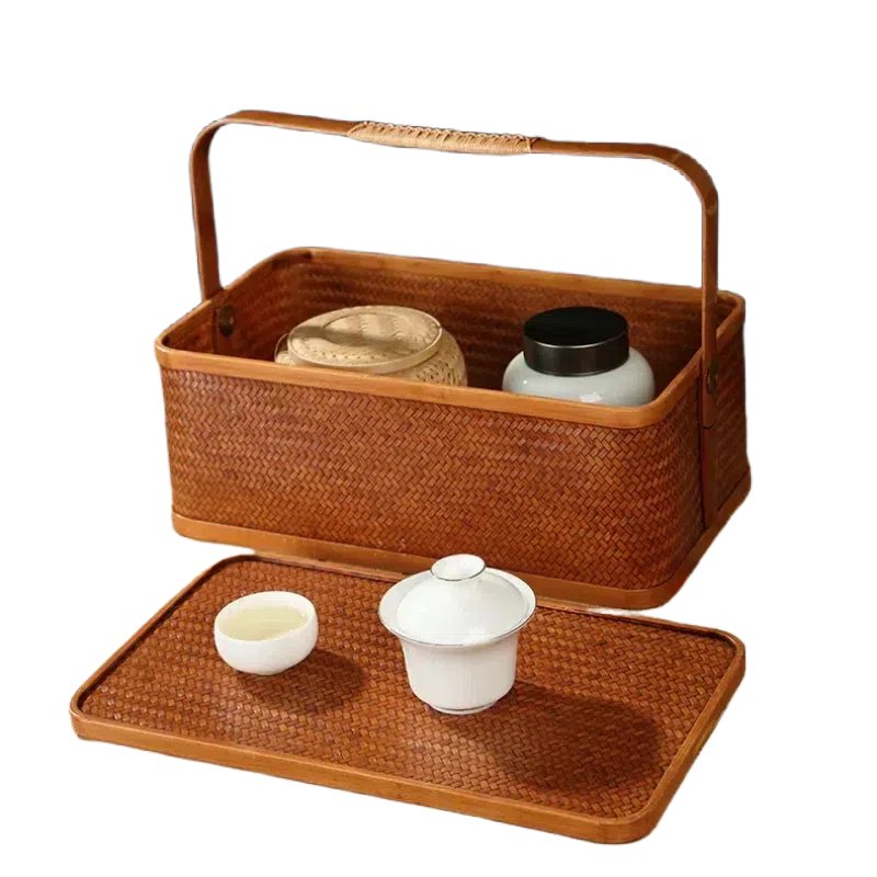 A woven rectangular basket with a handle, featuring a lid placed in front. On the lid are a white ceramic teacup and a lidded bowl. Inside the basket are two jars, one beige and one black.