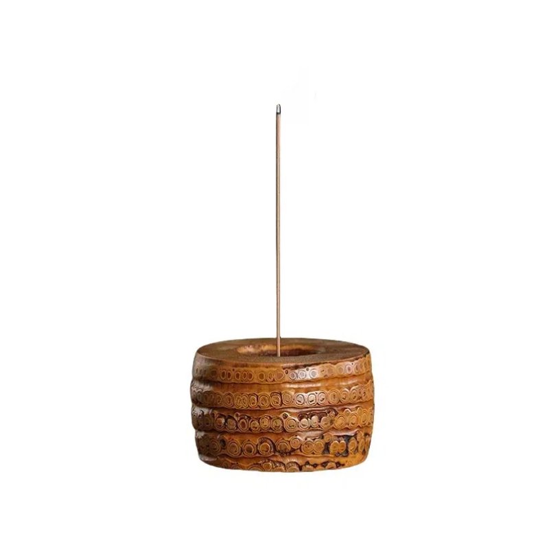 A carved wooden incense holder with intricate circular patterns, featuring an incense stick standing upright in the center. The holder has a cylindrical base and is set against a plain white background.