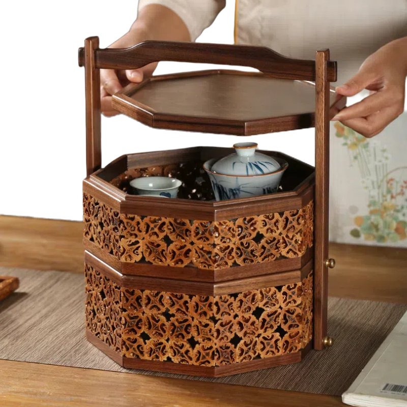 Someone arranges the New Chinese-style Walnut Craft Pastry Box Featured Image03, an intricately carved wooden food carrier with three tiers, on a table covered with a woven mat. The top tier holds blue and white porcelain bowls.